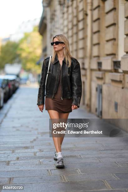 Natalia Verza wears black sunglasses from Prada, silver earrings, a gold chain pendant necklace, a dark brown shiny leather zipper coat, a brown silk...