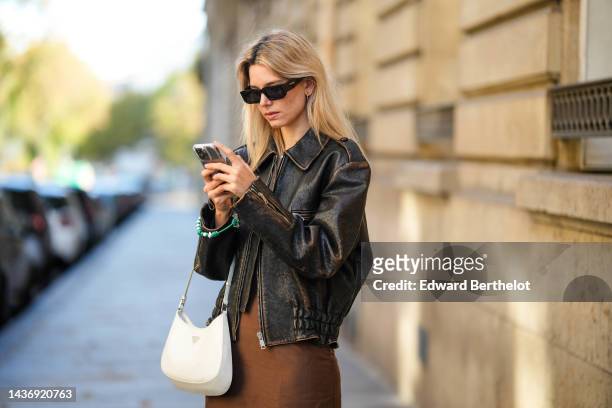 Natalia Verza wears black sunglasses from Prada, silver earrings, a gold chain pendant necklace, a dark brown shiny leather zipper coat, a brown silk...