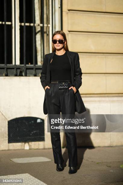 Diane Batoukina wears black sunglasses, a black tank-top, a black blazer jacket, a black shiny leather Chanel belt beg from Chanel, black large suit...