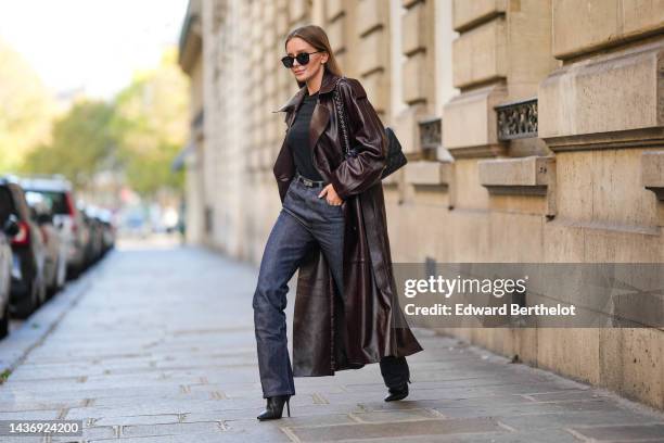 Diane Batoukina wears black sunglasses, a black t-shirt, a dark brown shiny leather long coat, a black shiny grained leather Kelly small belt from...