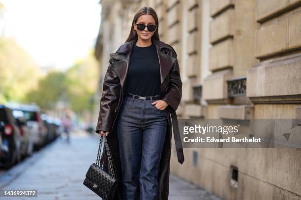 Diane Batoukina wears black sunglasses, a black t-shirt, a dark brown shiny leather long coat, a black shiny grained leather Kelly small belt from...