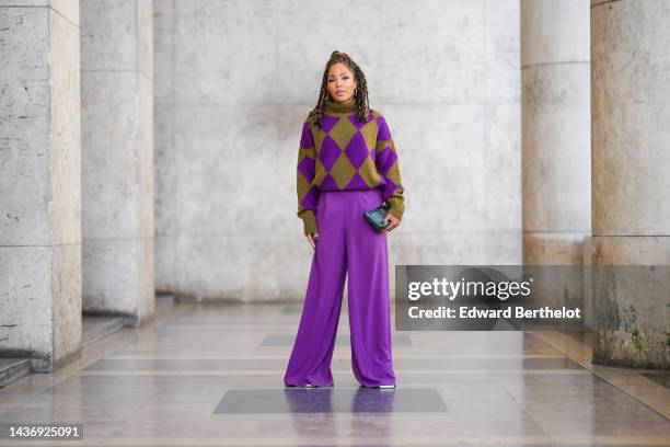 Ellie Delphine wears gold earrings, a khaki and purple checkered jacquard print pattern wool / turtleneck pullover, neon purple suit wide legs pants,...