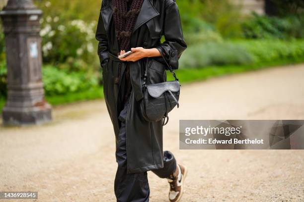 Guest wears a brown braided wool scarf, a black shiny leather belted long coat, black large puffy pants, a black shiny leather embossed Dior Oblique...
