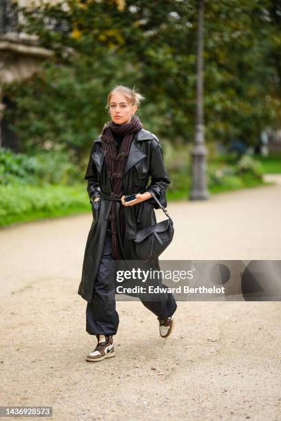 Guest wears silver earrings, a brown braided wool scarf, a black shiny leather belted long coat, black large puffy pants, a black shiny leather...