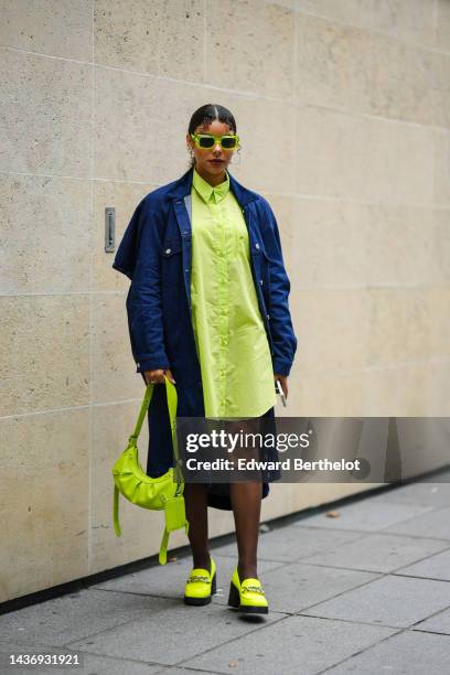 Guest wears neon yellow square sunglasses, gold and white pearls pendant earrings, a neon yellow striped print pattern shirt dress, a navy blue long...