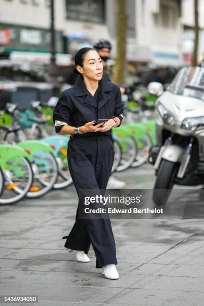 Guest wears diamond earrings, a black denim shirt jumpsuit, a silver large bracelet, silver rings, white matte leather block heels ankle boots ,...