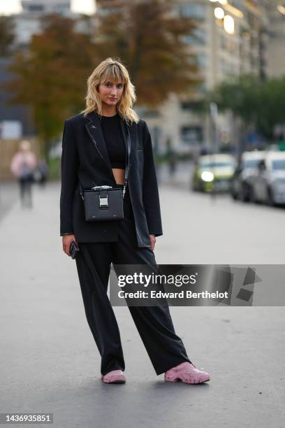 Guest wears a black cropped t-shirt, a black blazer jacket, a black shiny leather crossbody bag, black large suit pants, pale pink leather sneakers ,...