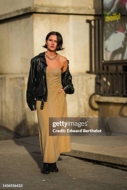 Guest wears a silver large chain necklace, a black shiny leather zipper biker jacket, a beige nailed / studded heart neck / long dress, black shiny...