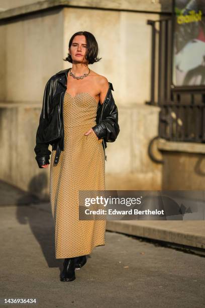 Guest wears a silver large chain necklace, a black shiny leather zipper biker jacket, a beige nailed / studded heart neck / long dress, black shiny...