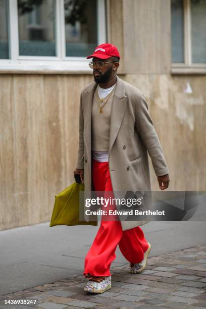 Guest wears a neon red cap, glasses, a white t-shirt, gold chain pendant necklaces, a beige wool pullover, a beige long wool coat, a yellow shiny...