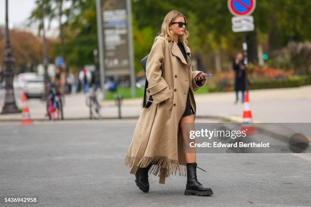 Guest wears black sunglasses, a black short dress, a black shiny leather shoulder bag, a beige fringed wool long coat, black shiny leather laces...