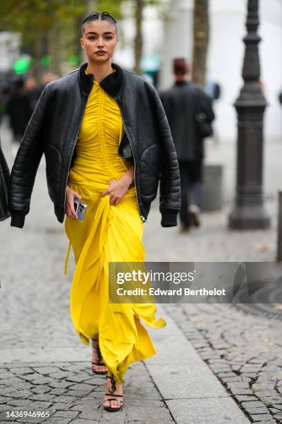 Guest wears a black shiny leather bomber coat, a yellow wrap / ruffled long dress, black shiny leather heels sandals , outside Stella McCartney,...