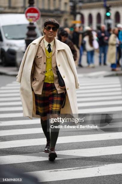 Guest wears a brown checkered print pattern felt wool beret, brown sunglasses, a pale yellow shirt, a black tie, a yellow wool buttoned cardigan, a...