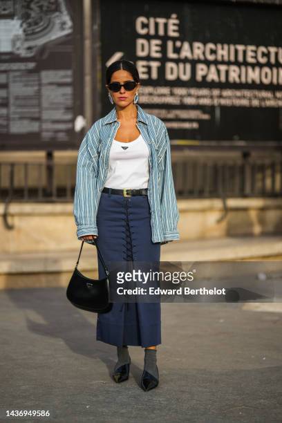Guest wears black sunglasses, blue earrings, a white tank-top from Prada, a blue and white striped print pattern shirt, a black shiny leather belt, a...