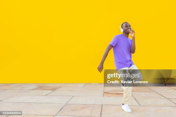 happy young african man with headphones and purple t-shirt dancing against yellow background - happiness and cheerfulness concept - boys dancing stock pictures, royalty-free photos & images