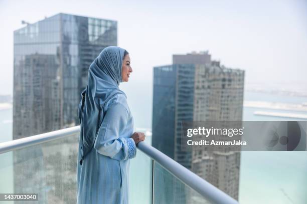 eine reife frau aus dem nahen osten beobachtet dubai von einem hohen balkon aus. sie ist ruhig und glücklich. fokus auf vordergrund - zurückhaltende kleidung stock-fotos und bilder