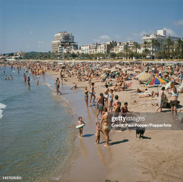 Visitors and holidaymakers sunbathe under parasol umbrellas and stand on the sandy Playa de Oro beach in the town of Sitges, a resort on the...