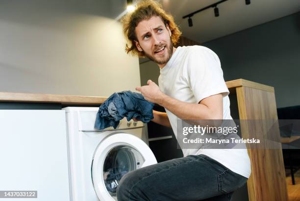 a young man is throwing things into the washing machine. domestic quarrel. problems of cleanliness in the house. cleaning company. vivid negative emotions of a man. - hair dryer stock photos et images de collection