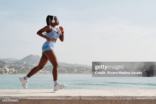 side view of a young black female athlete running on a stone wall with the coast in the background. - corriendo fotografías e imágenes de stock