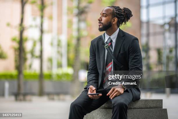 black salesman sitting on a concrete bench and using his smart phone - dreadlocks stock pictures, royalty-free photos & images
