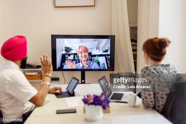 businesswoman seen on television screen greeting two colleagues in office during video call - board room background stock pictures, royalty-free photos & images