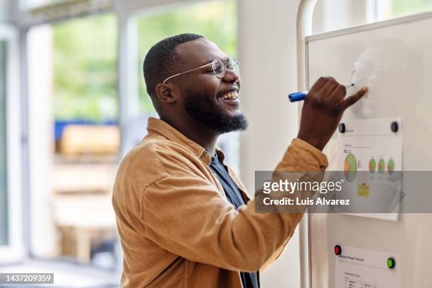 smiling african man writing on a whiteboard at a startup office - science collaboration stock pictures, royalty-free photos & images
