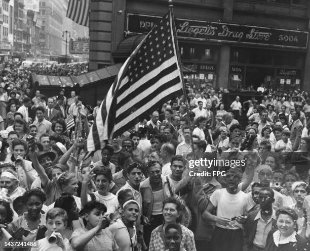 Crowds of people, one waving a stars-and-stripes flag, gather to celebrate Victory in Europe outside a branch of Liggett's drug store, in New York...