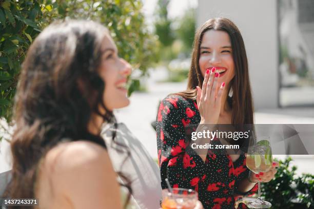 beautiful young women drinking cocktails, talking and laughing in café garden - mão na boca imagens e fotografias de stock