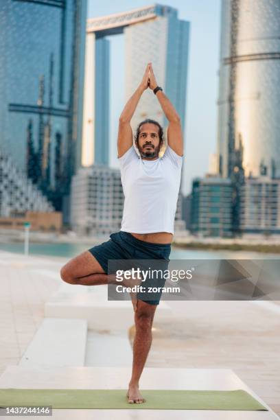 bearded man doing yoga, standing in tree pose on one leg with hands clasped on the pier in abu dhabi - yoga office arab stock pictures, royalty-free photos & images