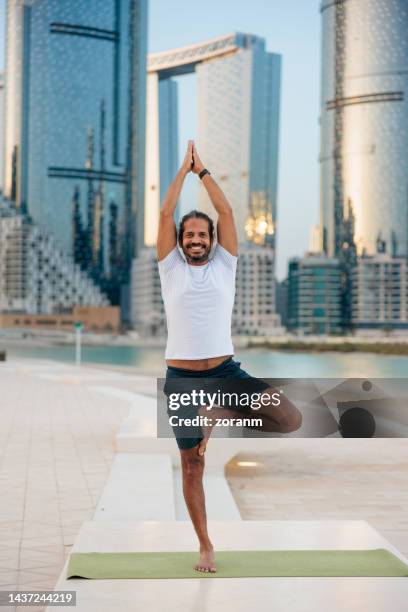 bearded man practicing yoga, standing in tree pose on one leg with hands clasped on the pier in abu dhabi - yoga office arab stock pictures, royalty-free photos & images