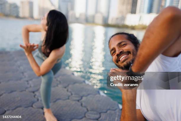 happy man and woman doing yoga by the sea, standing in lunge and turning to the side with hands clasped - yoga office arab stock pictures, royalty-free photos & images