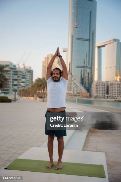 bearded man doing yoga, standing with arms raised and hands clasped on mat by the sea in abu dhabi - yoga office arab stock pictures, royalty-free photos & images