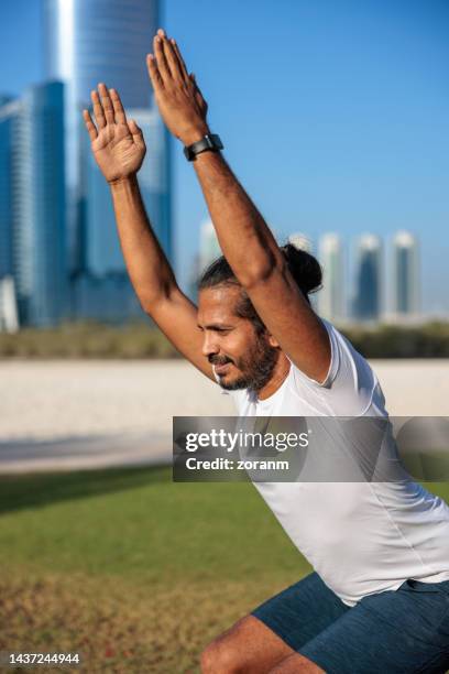 bearded man exercising on lawn in public park in abu dhabi squatting in chair pose - yoga office arab stock pictures, royalty-free photos & images