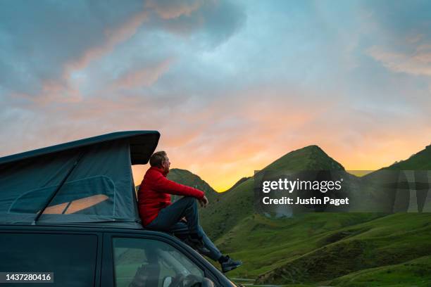 a mature man takes in the view of the beautiful sunrise in the mountains from the roof of his campervan - justin van groen stock pictures, royalty-free photos & images