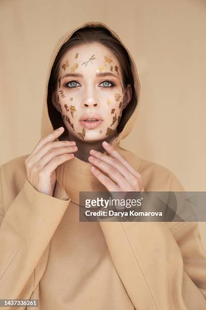 a young woman in a hood and beige suit stands on a beige background with gold decorative stickers on the face. the concept of islam woman, halloween party, beauty, makeup. - girl power stickers stock pictures, royalty-free photos & images