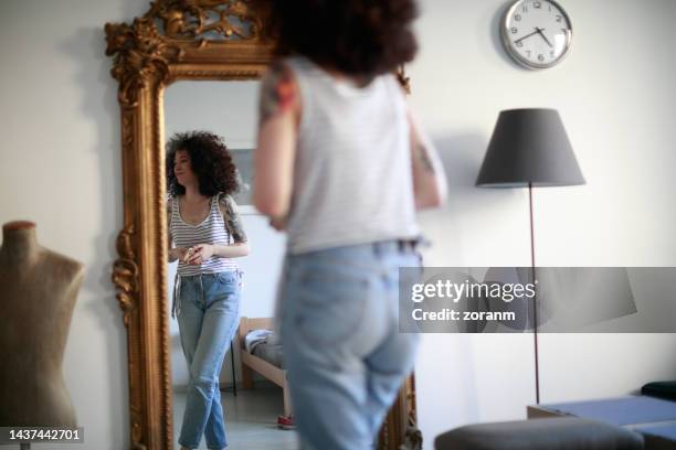 young tattooed woman with curly hair standing in front of full length mirror at home - woman full length mirror stock pictures, royalty-free photos & images