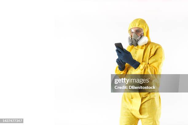 technician with yellow nuclear protection suit, mask and protective glasses consulting his mobile phone, on white background. concept of nuclear energy and pandemics. - schutzanzug stock-fotos und bilder