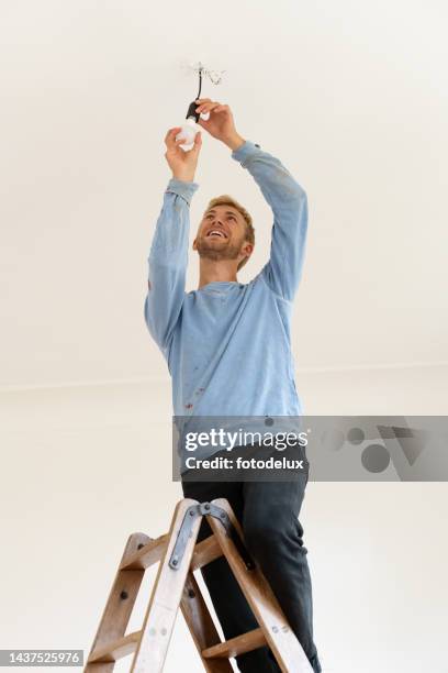young man installing electric bulb of living room ceiling - installing ceiling light fixture stock pictures, royalty-free photos & images