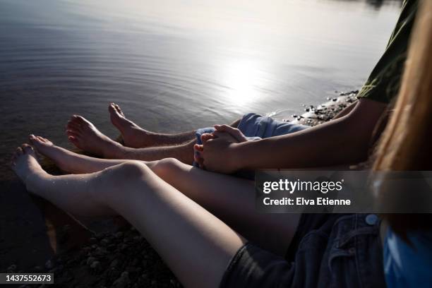 over the shoulder view of teenage couple holding hands together and sitting with feet dipped into the shallow waters at the shore of a lake in late afternoon autumn sunshine. - orilla del lago fotografías e imágenes de stock