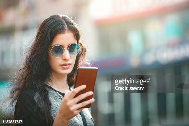 young woman uses a smartphone standing by the street. - lightskinned stock pictures, royalty-free photos & images