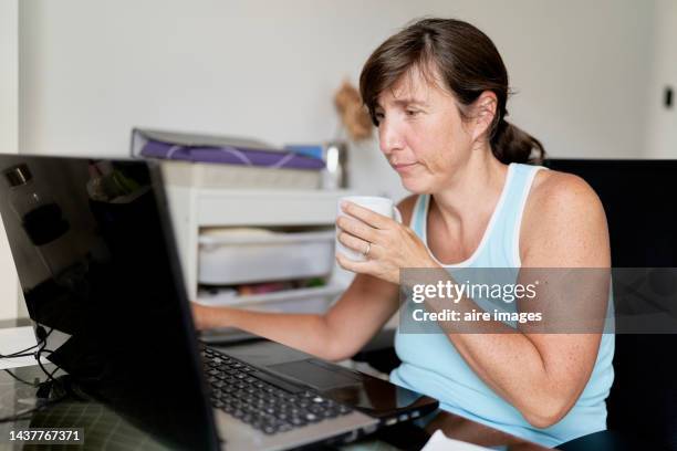 mature woman in casual clothes having a cup of coffee while working from a laptop in home - office concentration stock pictures, royalty-free photos & images