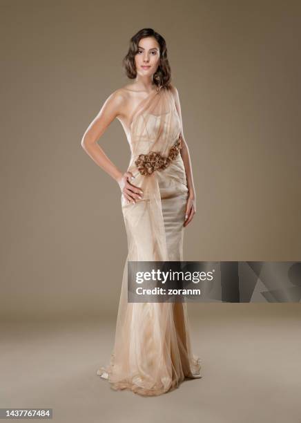 young brunette in champagne colored sleeveless maxi dress, hand on hip and looking at camera - champagnekleurig stockfoto's en -beelden