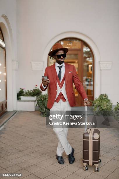 a black young man in a hat with dreadlocks and a brown suit and white trousers walks past the camera with a suitcase on wheels looks at the camera and smiles - white suit and tie stock pictures, royalty-free photos & images