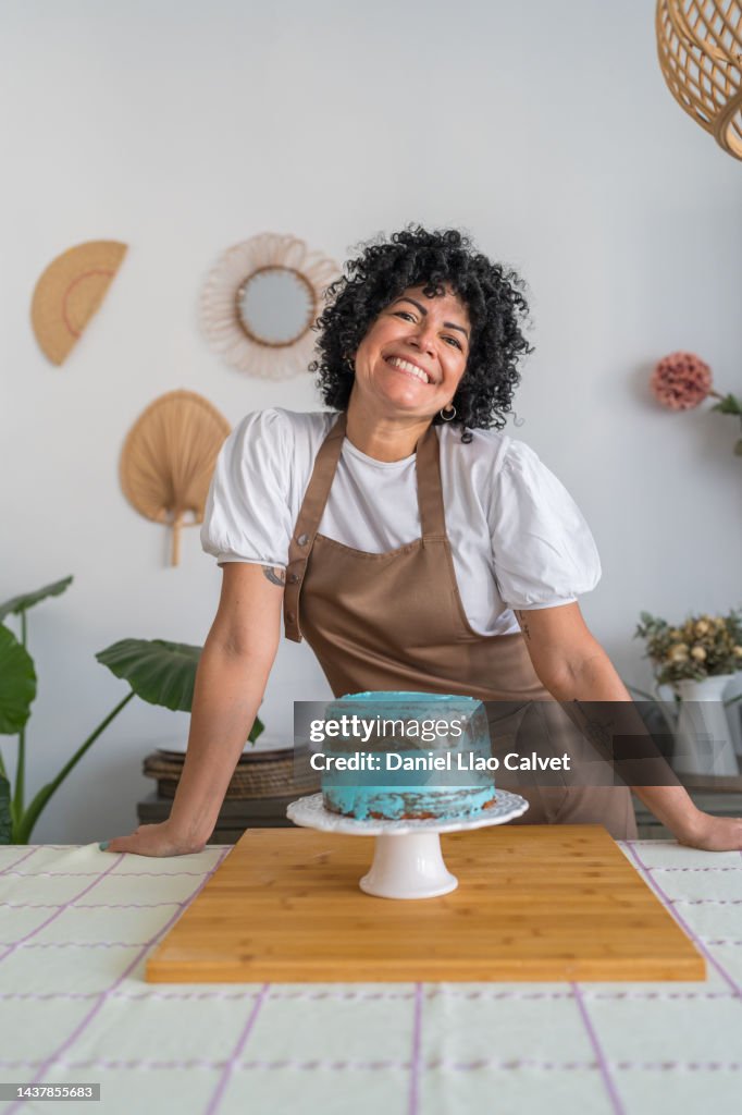 Positive mature ethnic female with curly hair smiling and looking at camera while standing at table with cake