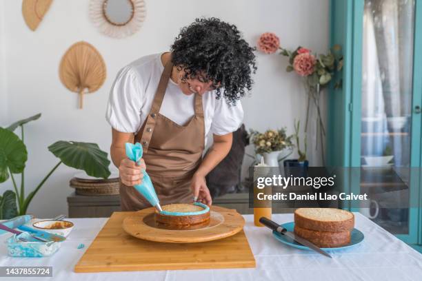 mature woman in brown apron decorate cake, squeezes out blue cream on a side of a cake - decorating a cake fotografías e imágenes de stock