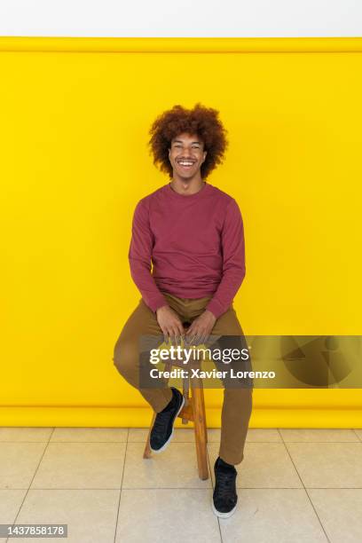 vertical portrait of happy black hipster young adult man smiling at camera sitting on stool in photo studio against yellow background - happy yello stock pictures, royalty-free photos & images