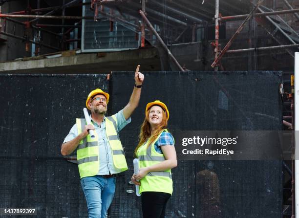 male and female construction worker colleagues smile happily as they look up at a building with scaffolding, the man pointing with his finger - vest stock pictures, royalty-free photos & images