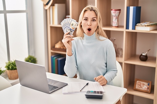 Young caucasian woman using laptop holding dollars banknotes scared and amazed with open mouth for surprise, disbelief face