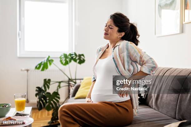 mulher jovem grávida que faz um rosto com as mãos no estômago enquanto está sentado em casa - muscular contraction - fotografias e filmes do acervo