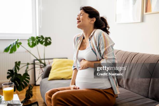 mulher jovem grávida em dor careta com as mãos no estômago enquanto sentada em casa - muscular contraction - fotografias e filmes do acervo